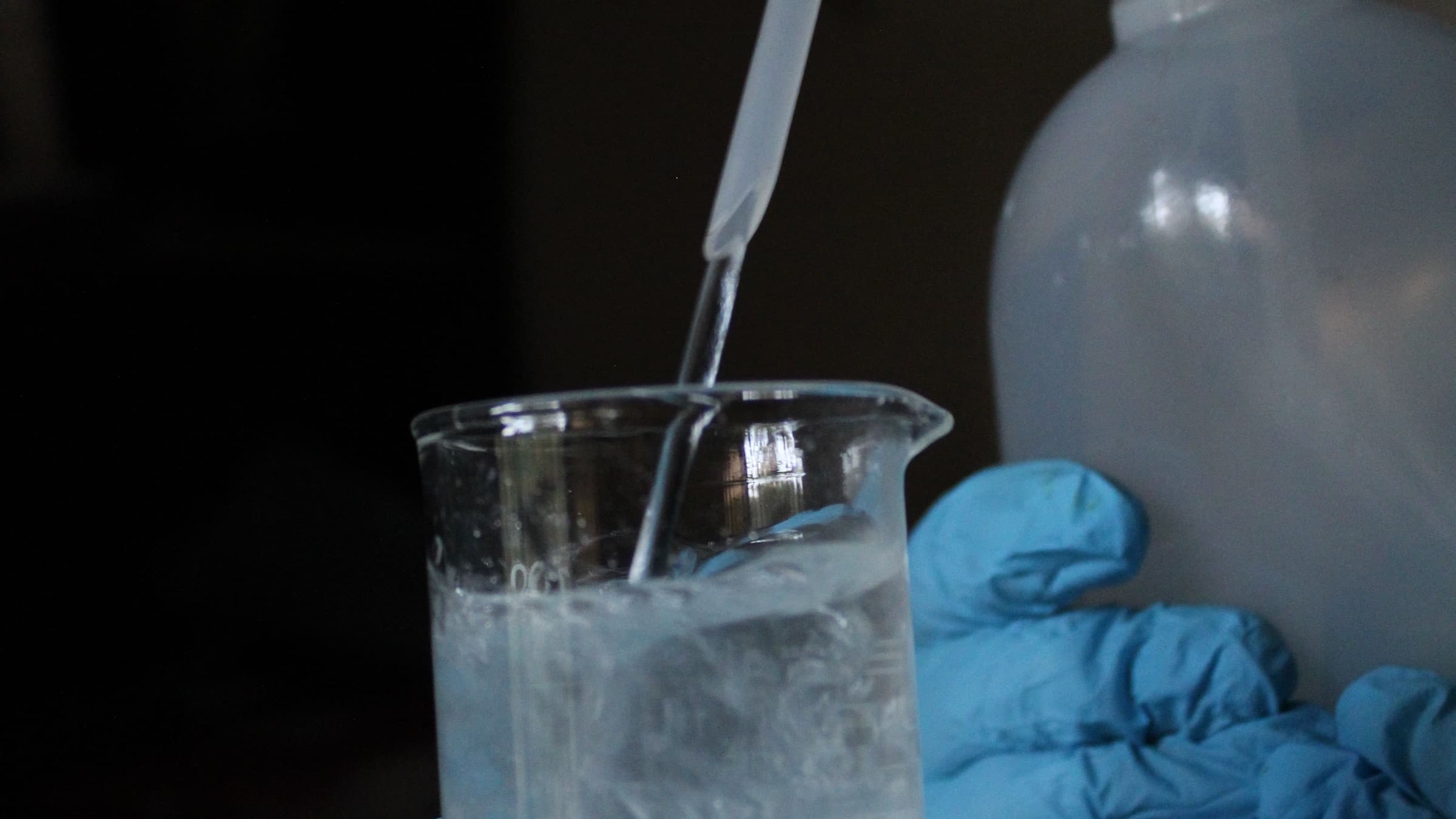 Gloved hand pipetting a clear liquid into a glass beaker