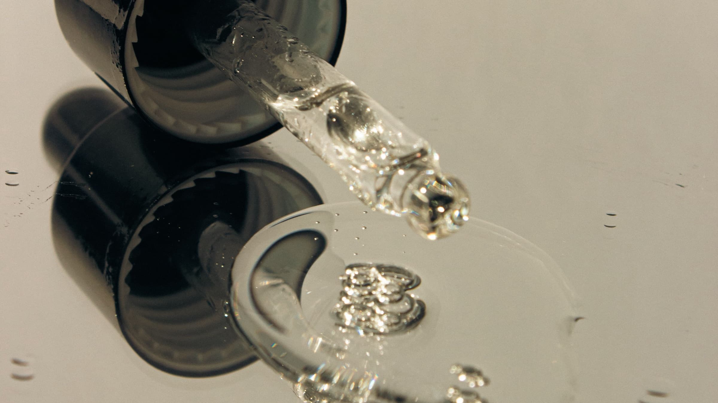 Glass dropper releasing a clear liquid onto a reflective surface